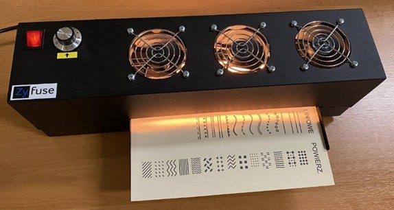 A black fuser for swell paper technique, equipped with three fans with metal guards and an analog temperature control knob. A tactile graphic with black patterns of lines, dots, and textures is emerging from the illuminated slot of the device. This process causes the printed graphics to become raised, creating a tactilely readable structure for people who are blind or have low vision.