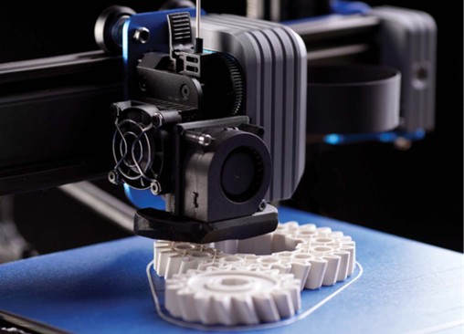 A complex 3D printer head with a black cooling fan and an exposed stepper motor is suspended just above a white pair of gears being printed. The mechanism is mounted on aluminum profiles. The entire scene is photographed in close-up from a slight front angle.