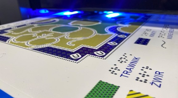 A colorful tactile graphic during UV printing, showing a garden plan with clearly raised textures and black Braille dots. On the white surface, the blue labels “LAWN,” “GRAVEL”, and “FLOWERS” are visible, accompanied by the corresponding tactile symbols in the legend. The entire scene is illuminated by the intense blue light of the ink-curing lamps, highlighting the three-dimensional structure of the printed details.